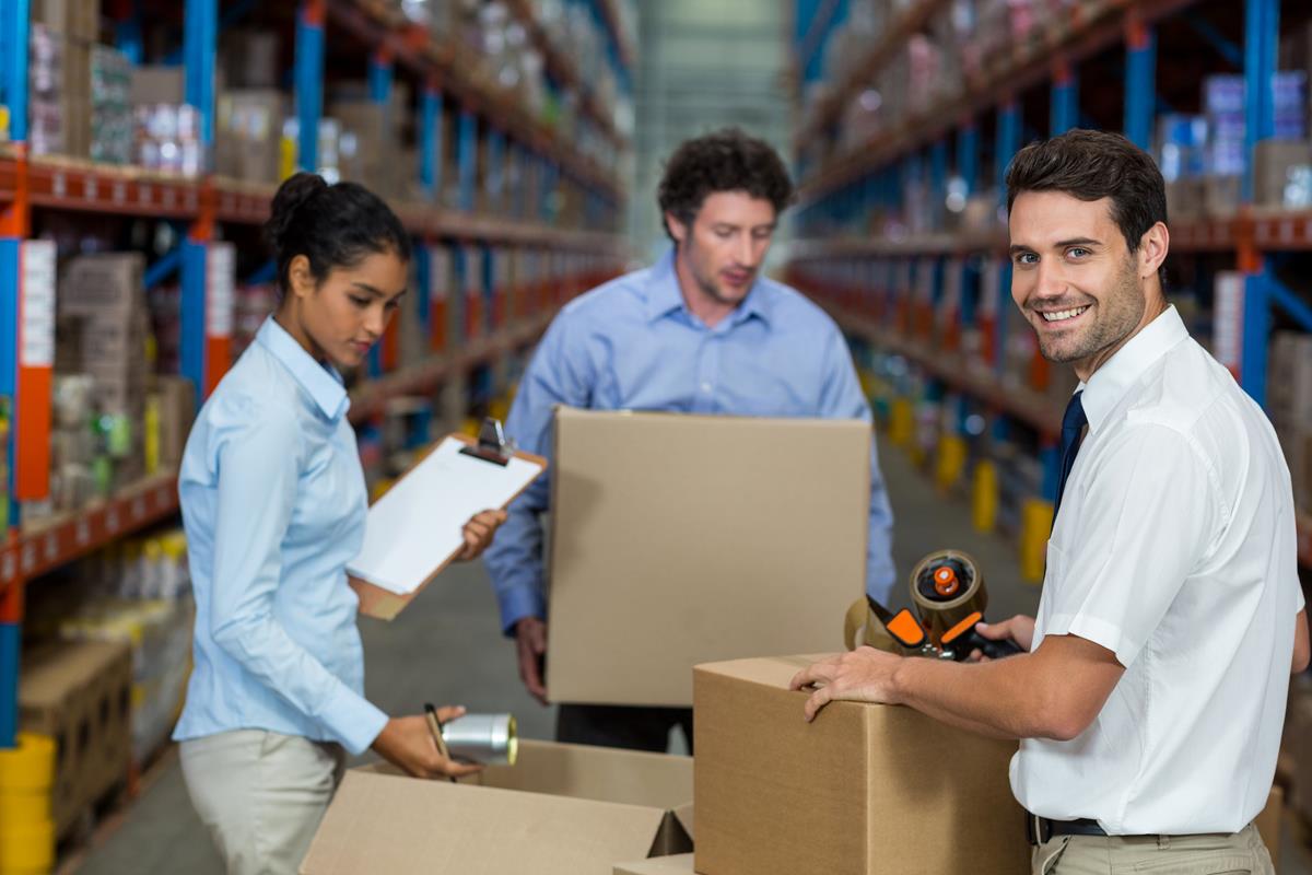 dois homens e uma mulher com uma pranxeta, coferindo mercadorias que estão dentro de uma caixa.